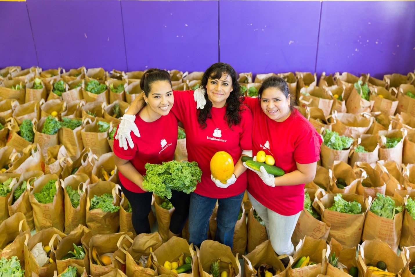 Three members posing in center of large grocery bags filled with vegetables.