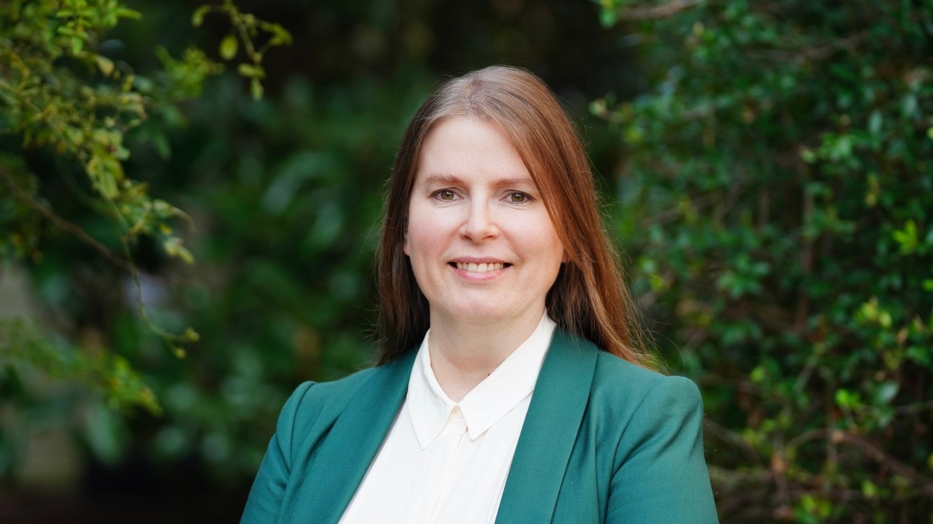 Woman wearing an emerald sport coat and white shirt smiling at camera, standing in front of trees and foliage.