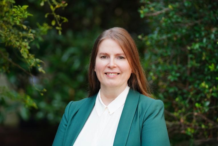 Woman in green sports coat smiling and standing in front of green foliage