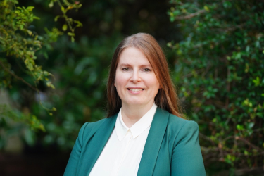 Woman in green sports coat smiling and standing in front of green foliage