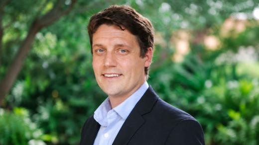 Man in a dark blazer and light blue shirt smiling outdoors against a leafy green background.