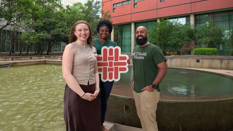 Three students standing in front of fountain holding a school logo sign.