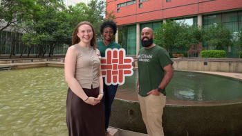 Three students standing in front of fountain holding a school logo sign.